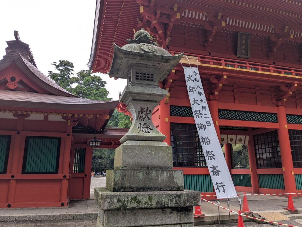 石灯籠の奥に朱色の楼門と祭事の白い幟が見える境内